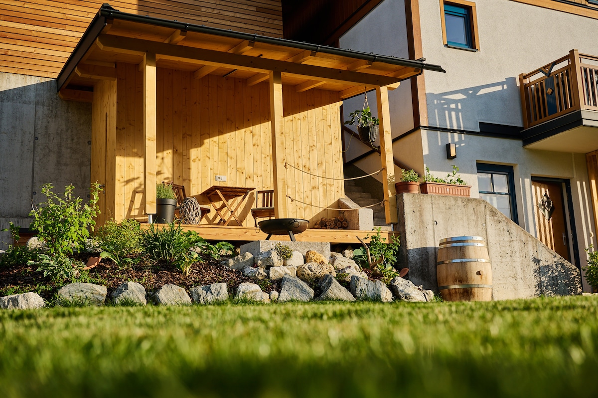 A wooden terrace is showcased in the image, featuring a rustic seating area and a small campfire setup. Lush green grass surrounds the space, with carefully arranged stones and plants adding natural elements. The building's architecture offers a harmonious blend of wood and stone.
