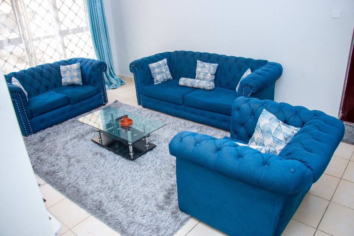 A living area is furnished with a plush blue sofa set, featuring tufted details and decorative cushions. A glass coffee table sits at the center, resting on a soft, light gray area rug. Natural light enters through large windows, accompanied by sheer curtains.