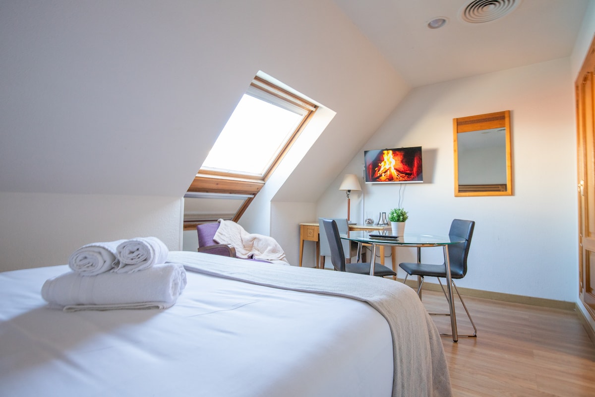 A spacious room features a comfortable bed with neatly arranged towels. A dining setup is present with a table and two chairs. Natural light enters through a slanted window, and a wall-mounted TV displays a serene fireplace image. A mirror reflects the room's brightness.