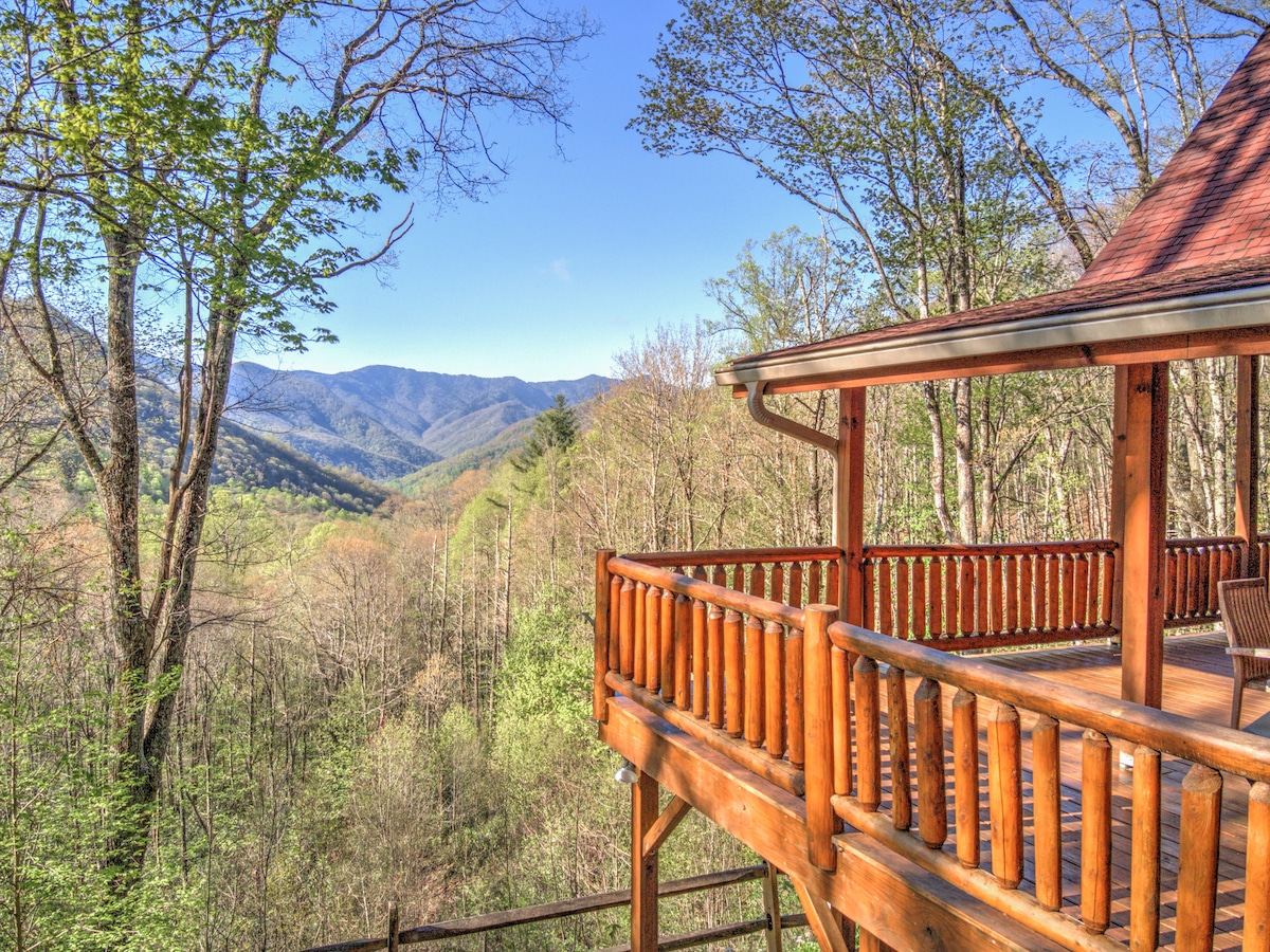A wooden deck with rustic log railings offers a scenic view of rolling mountains and dense trees. The surrounding landscape is marked by a variety of greens, indicating a rich natural environment. Clear blue skies stretch above, enhancing the outdoor experience.