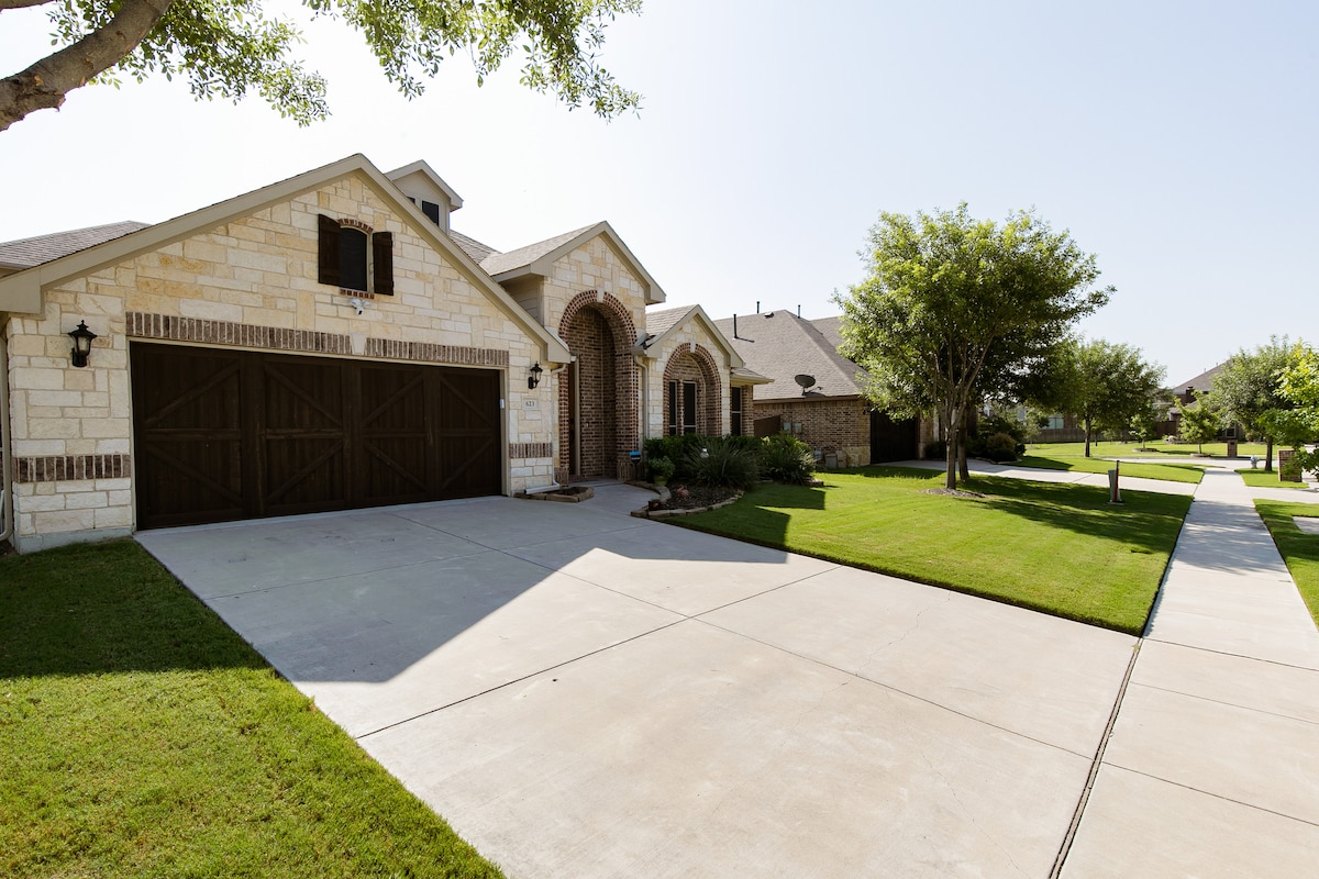 The driveway leads to a spacious garage with double doors, surrounded by well-maintained lawn and landscaped borders. Sidewalks extend invitingly along the property, with trees providing gentle shade on a clear day.
