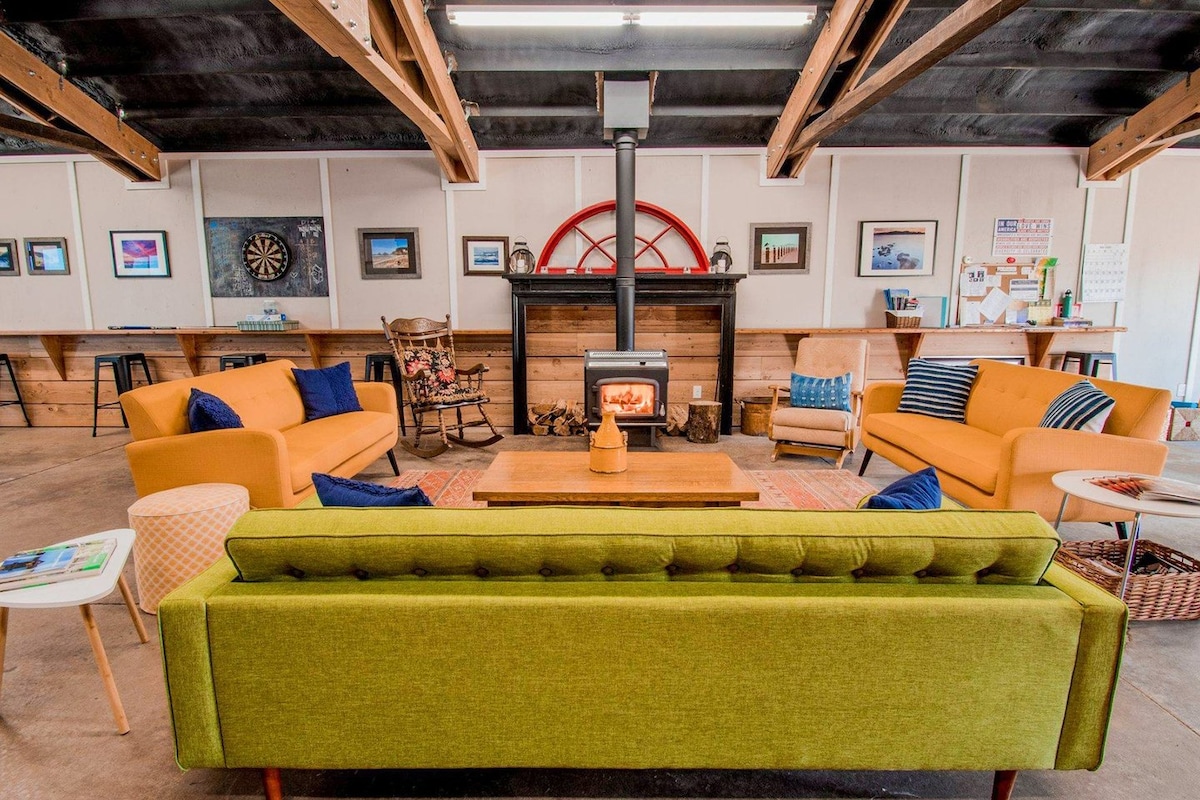A spacious living area features a vibrant green sofa and warm yellow chairs arranged around a rustic wooden coffee table. A cozy fireplace is positioned against a wooden wall, complemented by framed artwork. A soft rug adds texture to the concrete floor.