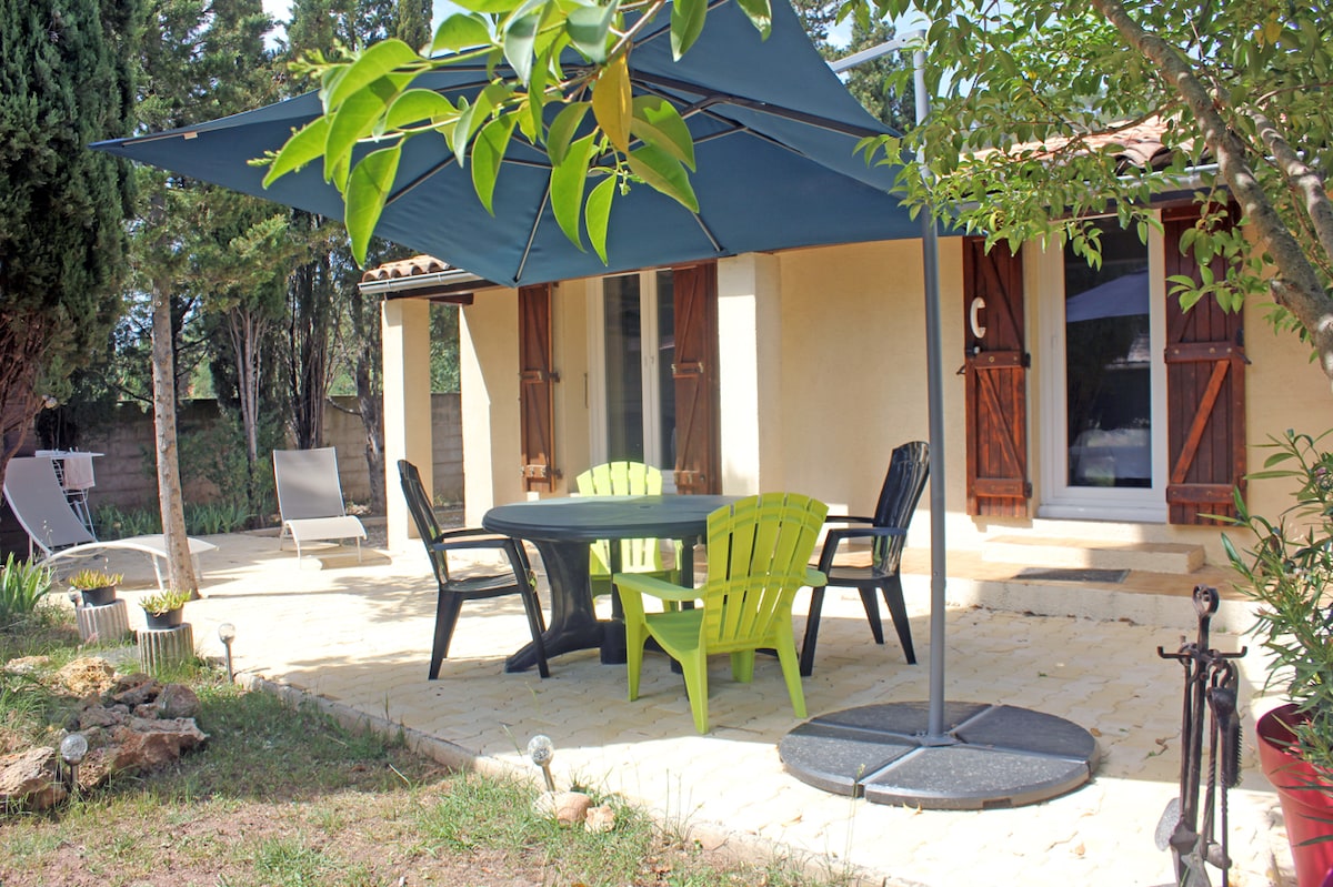 A covered outdoor patio features a rectangular dining table surrounded by four chairs in varying colors. Sunshade fabric extends overhead, providing shade from sunlight. Lush greenery surrounds the area, with a simple pathway leading to the residence in the background.