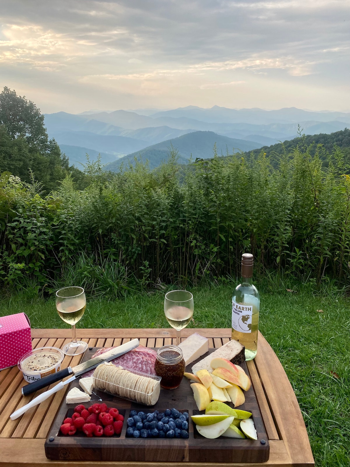 A wooden table is set outdoors overlooking a scenic mountain view. Two glasses of white wine sit beside a platter featuring assorted cheeses, fruits like apples and berries, and a jar of jam. The sky is lightly cloudy, creating a serene backdrop.