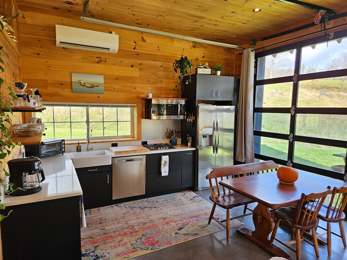 An open kitchen area features modern appliances, including a stainless steel refrigerator and four-burner stovetop. Light-colored wooden walls complement dark cabinetry. A rustic wooden dining table surrounded by chairs is positioned near large windows, allowing natural light to illuminate the space. A decorative pumpkin adds a seasonal touch.