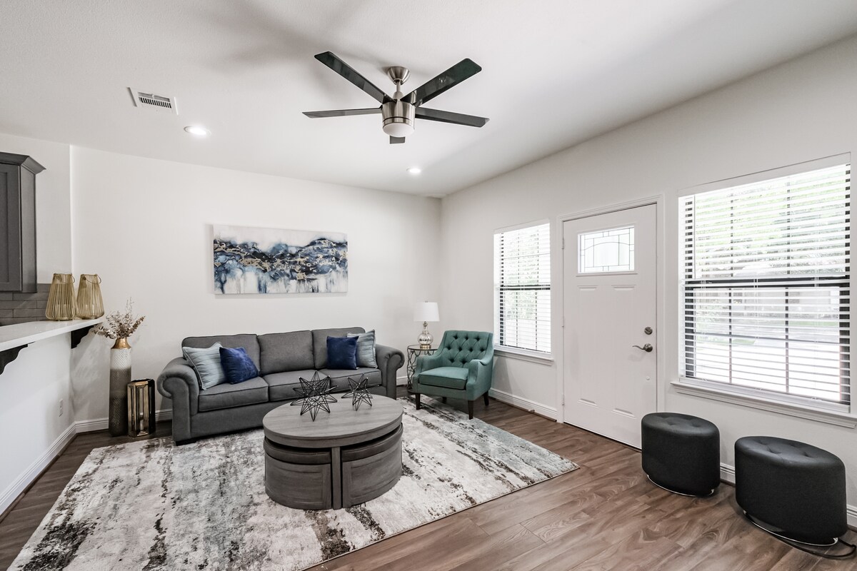 A spacious living area features a gray sofa paired with a green accent chair. A round coffee table is centered on a large area rug, complementing the wood flooring. Natural light flows through the windows, which are adorned with blinds for light control.