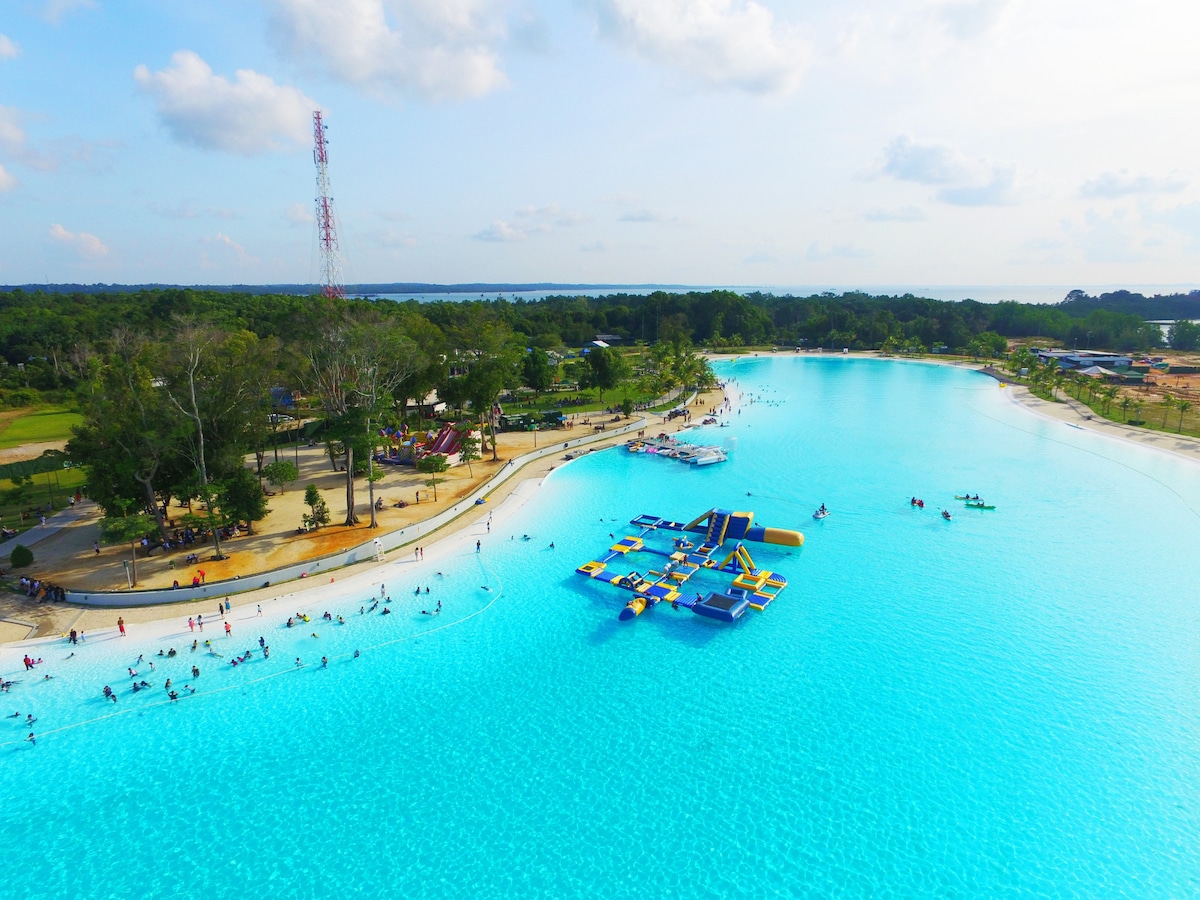 The expansive Crystal Lagoon is shown, featuring vibrant turquoise waters surrounded by sandy beaches and lush greenery. Floating inflatables are visible in the water, and various recreational activities can be spotted along the shoreline. Visitors can be seen enjoying the lagoon under a partly cloudy sky.