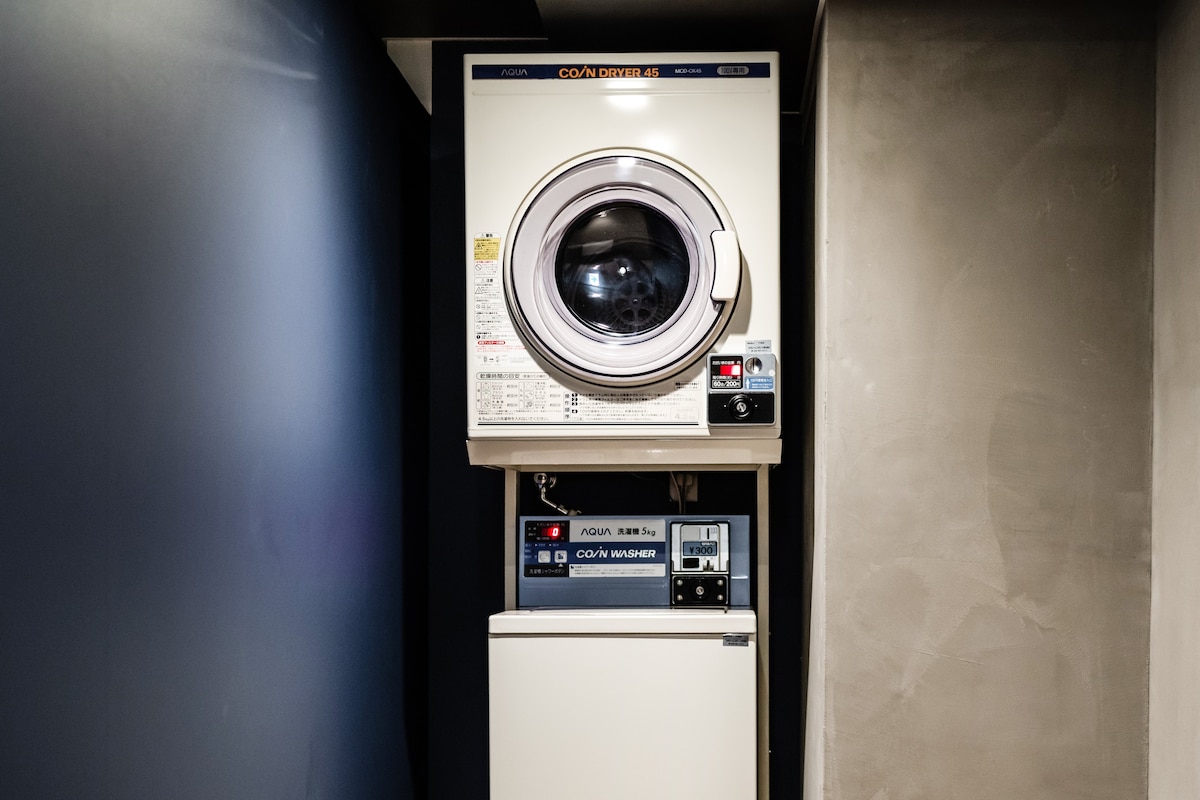 A stackable washer and dryer unit is positioned against a neutral-colored wall. The laundry appliances are compact and feature front-loading designs, with a digital display panel for convenient operation.