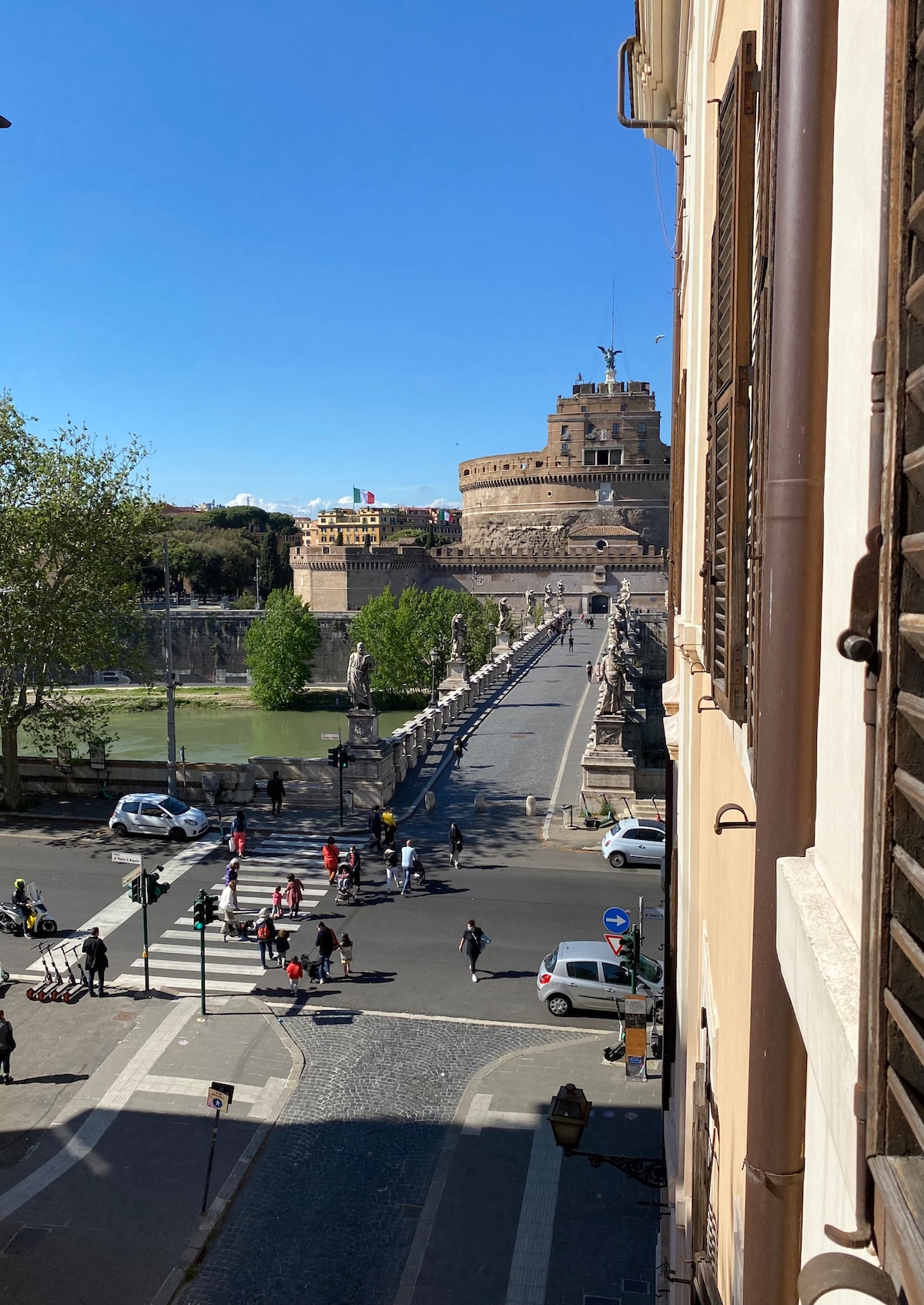 A view from a window reveals the serene landscape of a historic street. Castel Sant'Angelo is visible in the background, surrounded by greenery and blue skies. People can be seen walking across the stone bridge, emphasizing the lively atmosphere of the area.