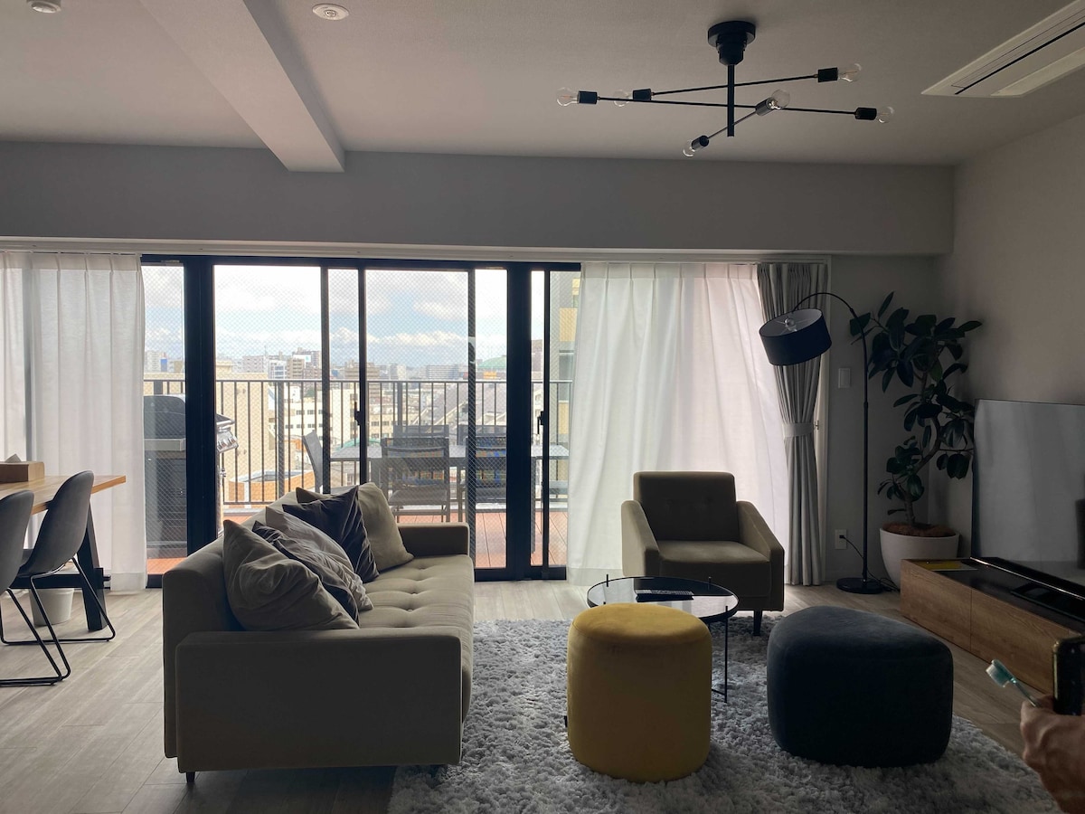 A modern living space features a comfortable sofa and colorful poufs arranged on a soft rug. Large glass doors lead to a balcony with city views, while natural light fills the room through sheer curtains. A sleek dining area and potted plant create a welcoming ambiance.