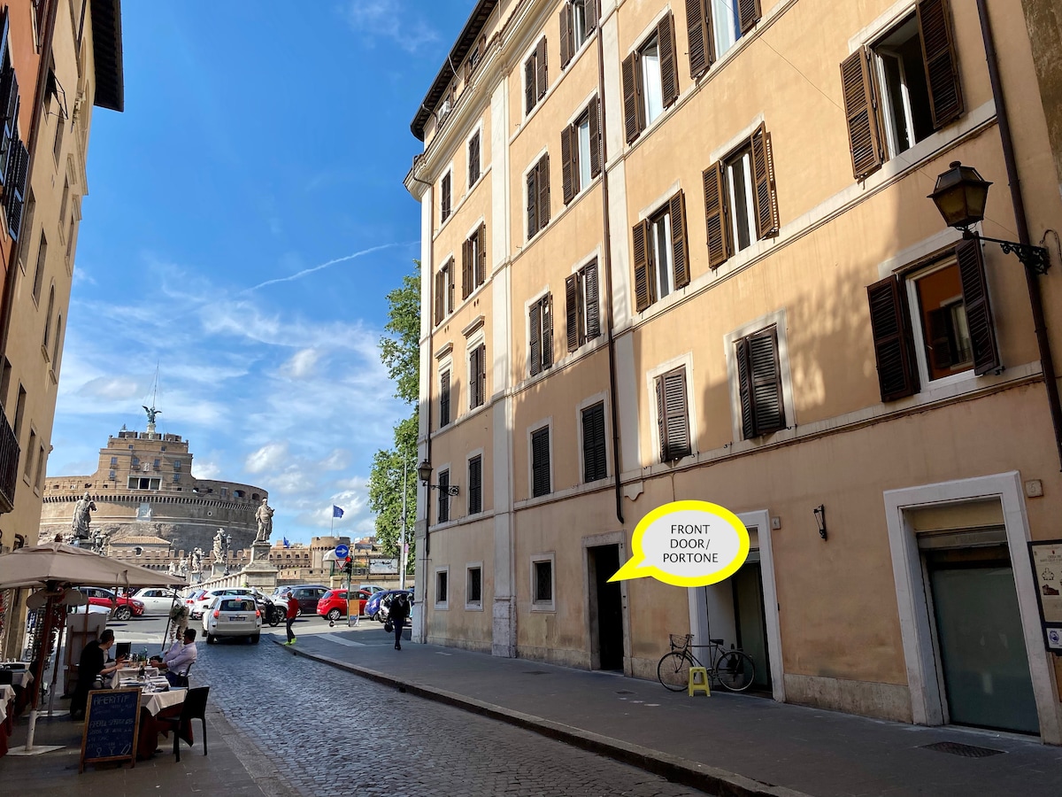 The exterior of the property is shown, featuring a multi-story building with traditional Roman architecture. Large windows with wooden shutters are present. A street scene includes bicycles parked near the entrance and a glimpse of Castel Sant'Angelo in the background.