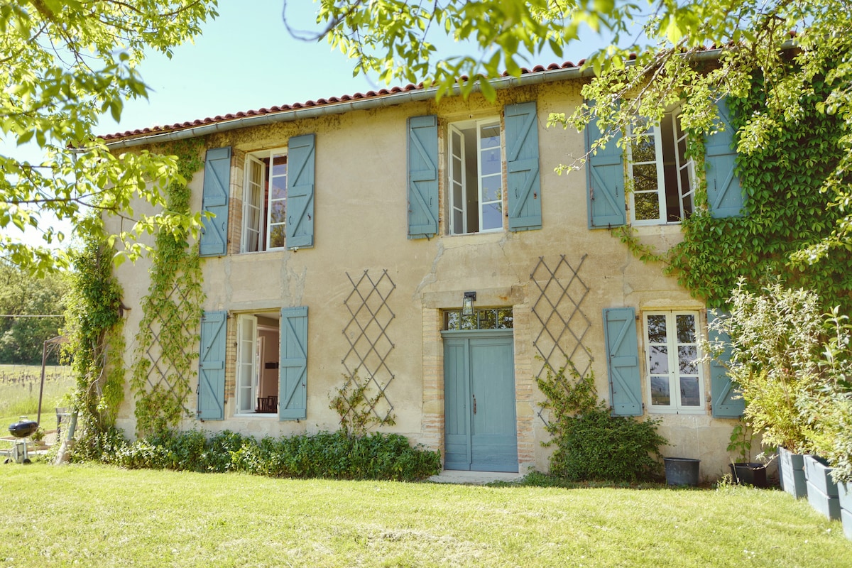 A charming two-story building features light blue shutters and a wooden front door, surrounded by greenery. The exterior showcases a mixture of stone and stucco, with climbing vines adding texture. A well-maintained garden area is visible, providing a serene outdoor space.