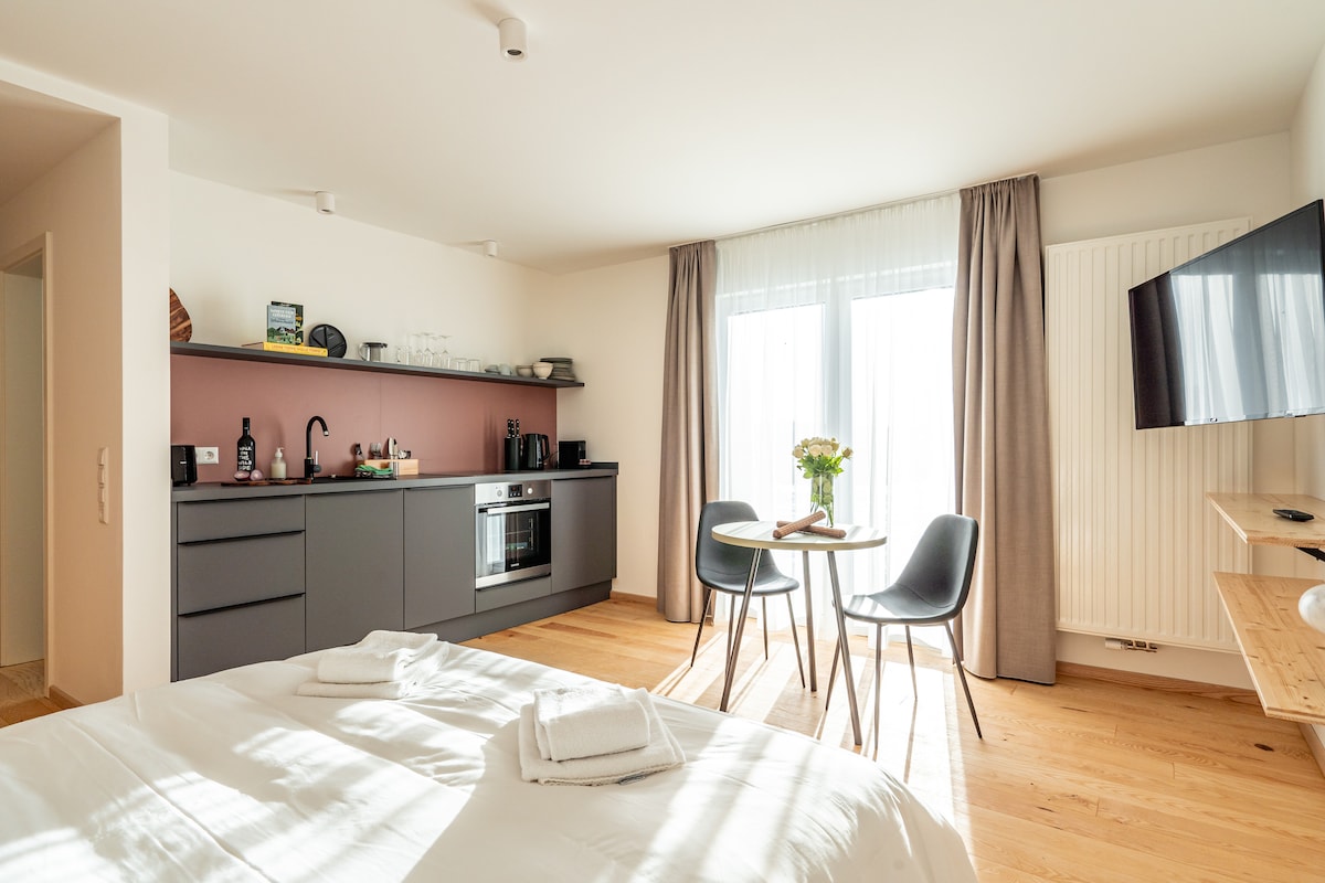 A well-appointed studio features a cozy sleeping area with a spacious bed. A kitchenette is visible, alongside a dining corner with a small table and two chairs. Natural light floods through large windows, complemented by neutral curtains, creating a bright and inviting space.