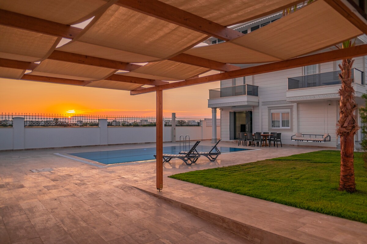A spacious outdoor seating area is shaded by a wooden structure, overlooking a private pool and lush green lawn. The sunset casts a warm glow across the scene, with the villa visible in the background and comfortable loungers arranged nearby.
