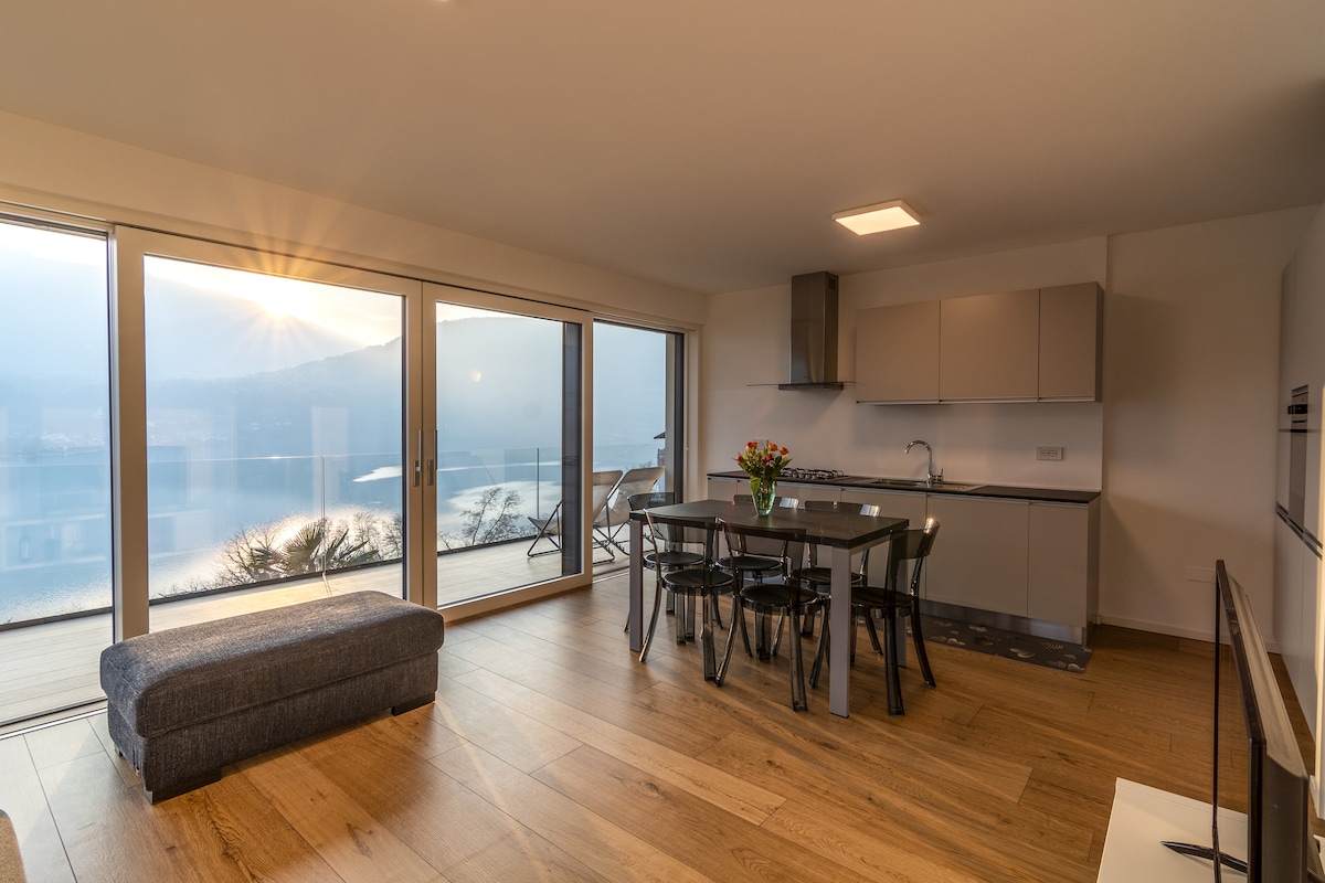 A modern kitchen area is featured, with sleek cabinetry and a dining table surrounded by black chairs. Large glass doors provide views of the lake and surrounding landscape, allowing natural light to brighten the space. A cozy seating option is positioned nearby.