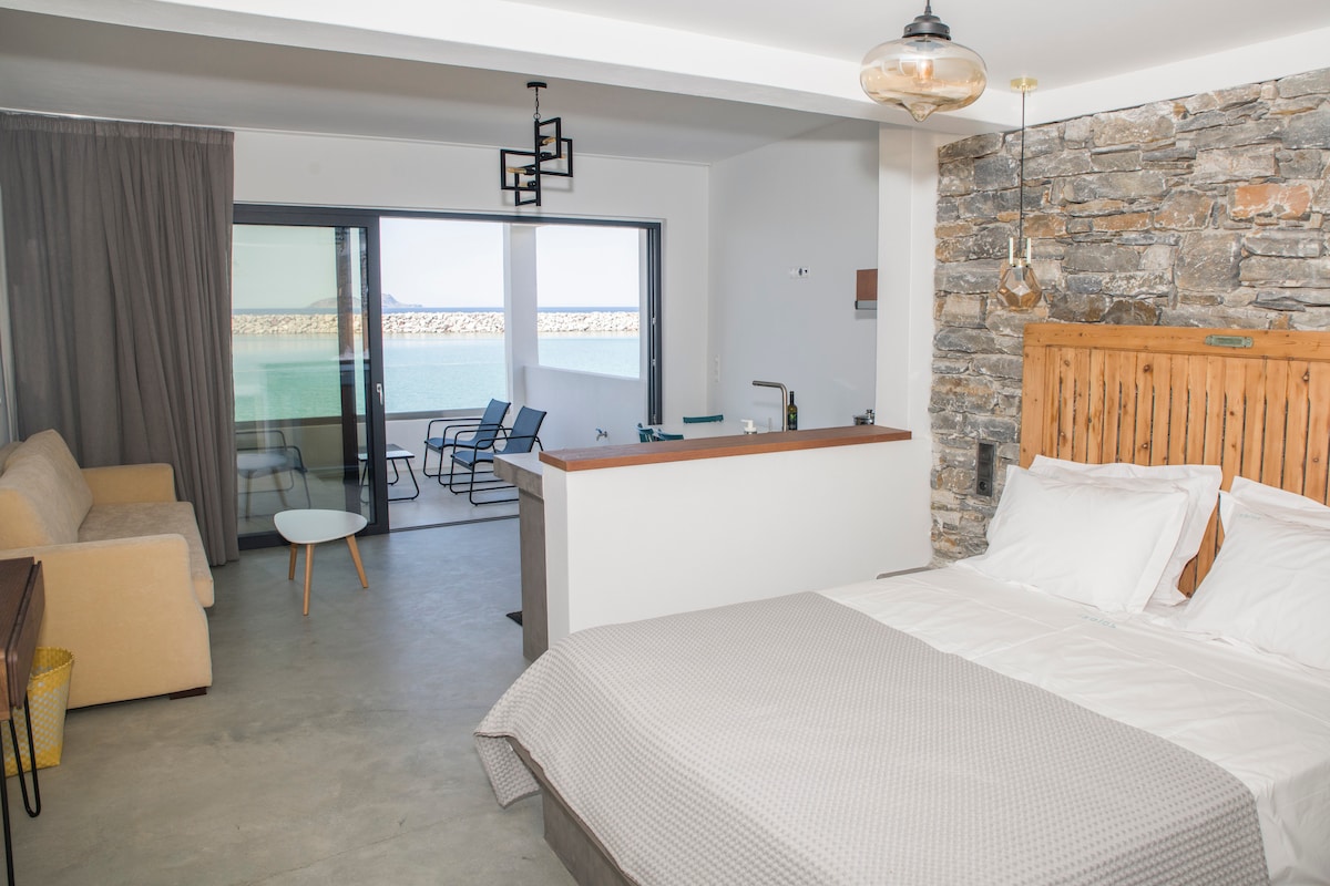The studio features a spacious sleeping area with a large double bed and a sofa that can convert into a bed. Natural light enters through sliding glass doors, revealing views of the sea. A kitchenette is visible, providing essential appliances for meal preparation.