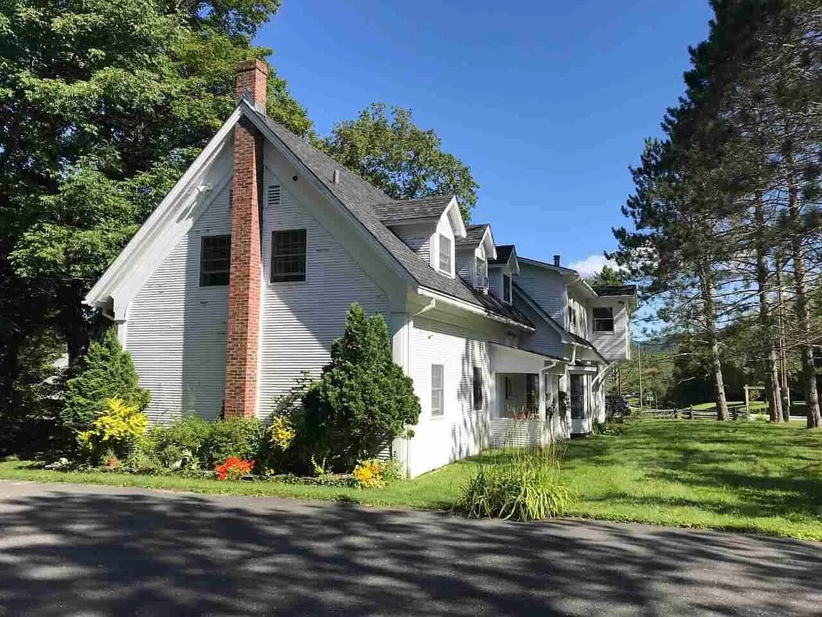 The exterior of the farmhouse features a white facade with a brick chimney. Lush greenery and blooming flowers surround the building, with a well-maintained lawn extending outwards. The building's pitched roofs and multiple windows reflect a classic architectural style, set against a clear blue sky.