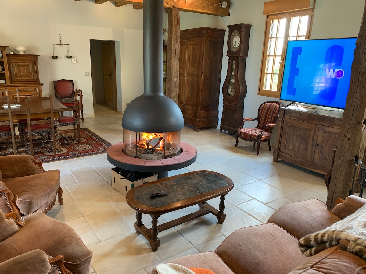 A communal living area features a centrally located modern fireplace with a circular design. Comfortable seating is arranged around a low coffee table, complemented by wooden cabinetry and a vintage clock. Light enters through windows adorned with simple treatments, enhancing the space's warmth.