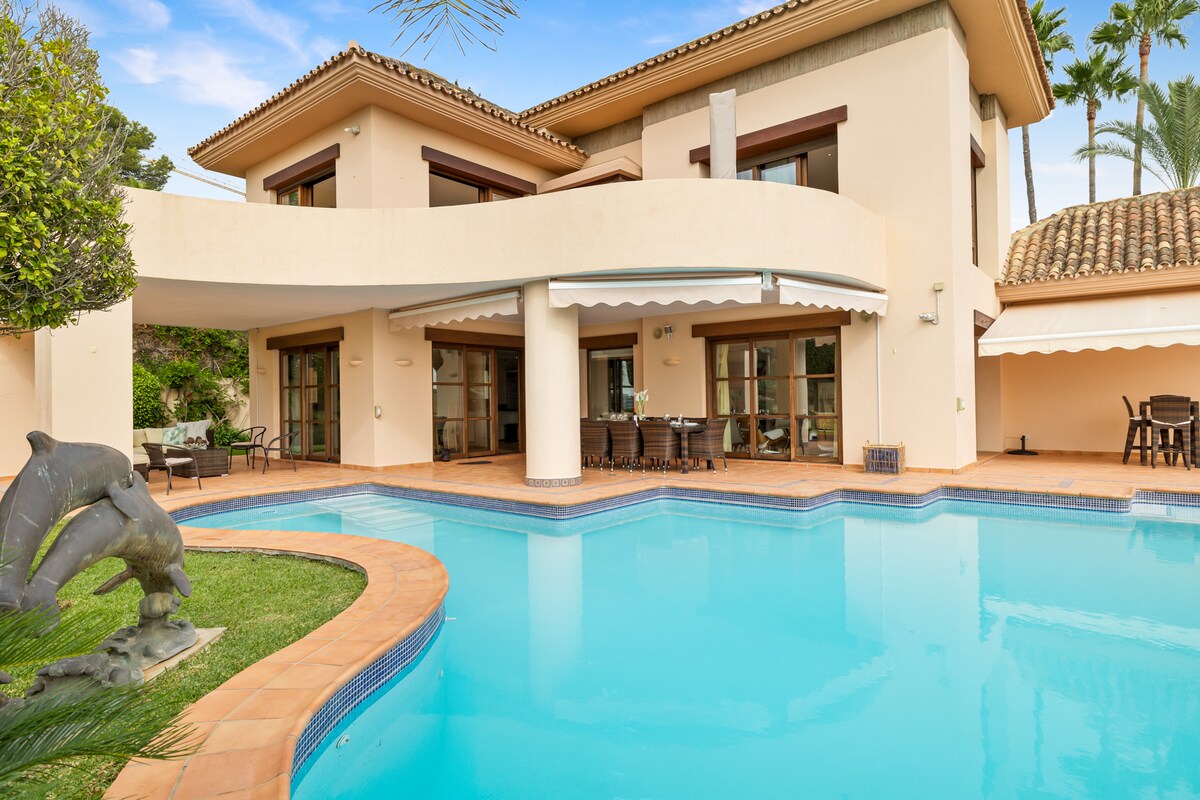 The exterior of the villa features a spacious swimming pool with clear blue water, surrounded by a tiled patio. Sliding glass doors provide access to dining and seating areas. Lush greenery and palm trees contribute to the serene atmosphere of the outdoor space.