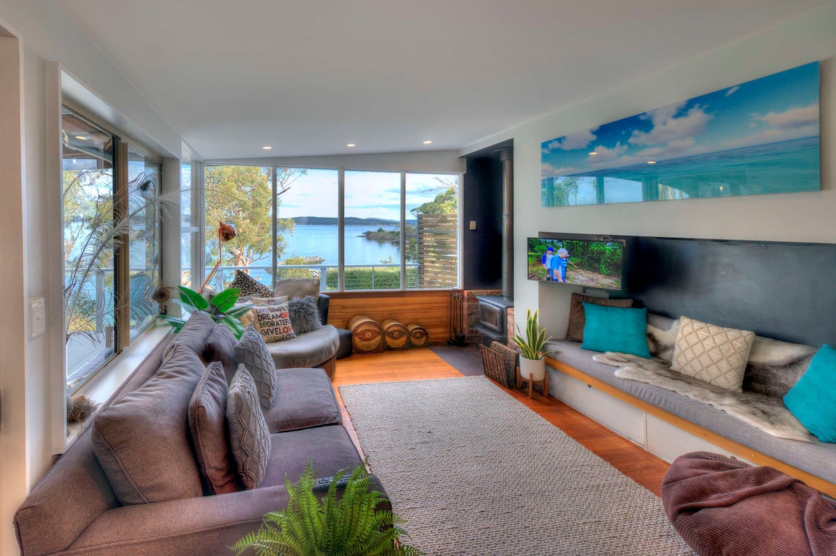 A comfortable lounge area features a sectional sofa and a funky round couch, inviting relaxation. Large windows provide expansive views of the ocean and surrounding landscape. A wall-mounted television and decorative cushions add to the cozy ambiance, while natural light fills the space.