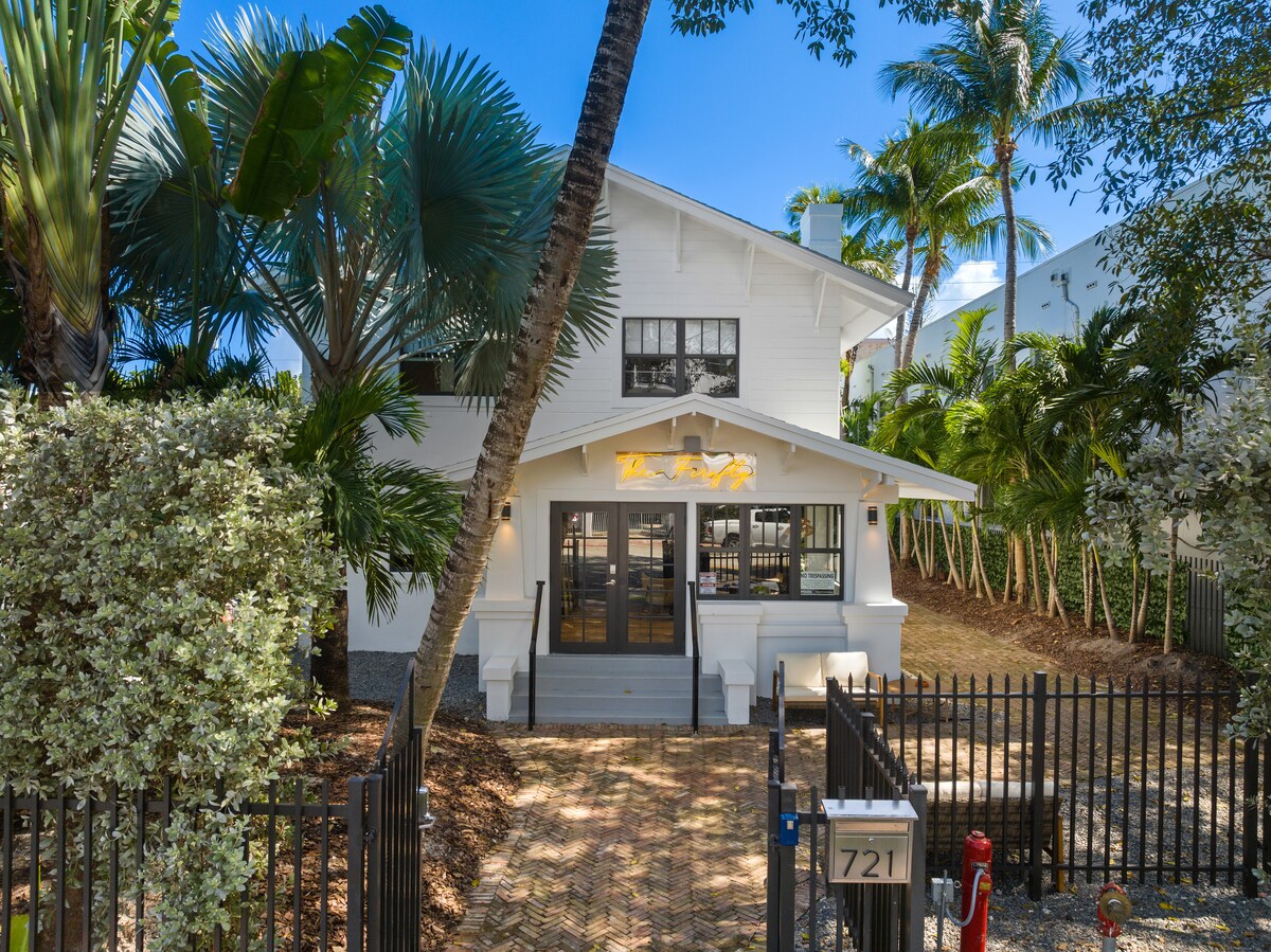 A charming two-story house is nestled among lush palm trees, featuring a welcoming front porch with large glass doors. A paved pathway leads to a gated entrance, while tropical foliage surrounds the property, enhancing the serene atmosphere of the setting.