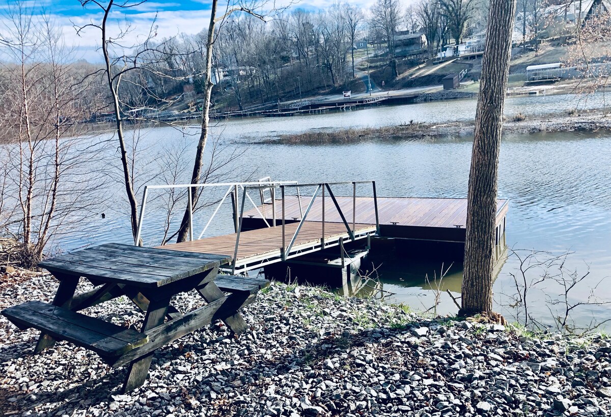 A wooden picnic table is positioned on rocky terrain near a calm lake, offering a serene view of the water and surrounding trees. A private dock extends into the lake, providing an accessible spot for water activities.