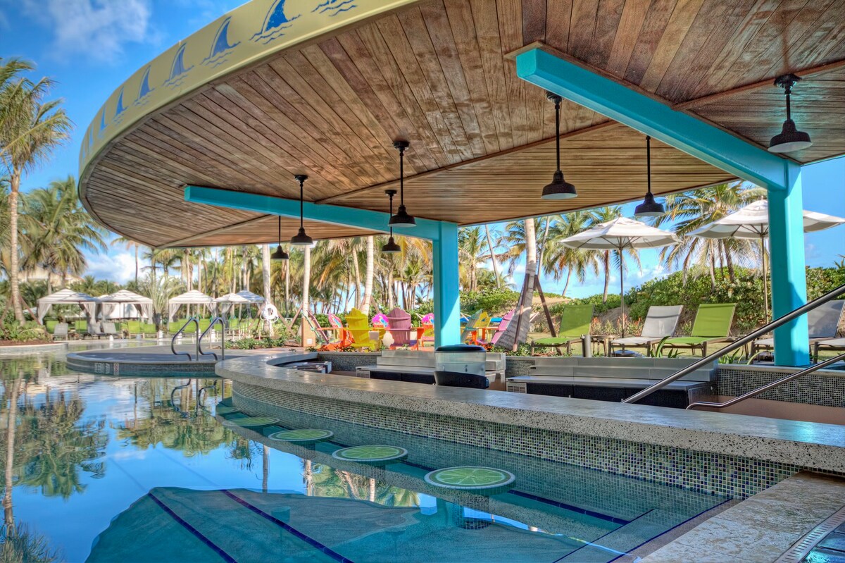 A circular pool bar with a wooden ceiling features stylish hanging lights. Brightly colored lounge chairs and umbrellas surround the pool, set against a backdrop of lush greenery and palm trees. The clear water reflects the blue sky and creates a relaxing ambiance.
