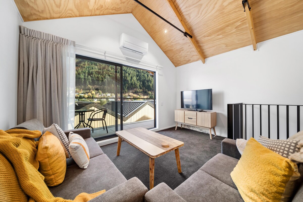 A comfortable living area features a modern design with a soft grey couch adorned with yellow and patterned throw pillows. A wooden coffee table is positioned in front. Large sliding doors offer views of the outdoors, complemented by a balcony and natural light filling the space.
