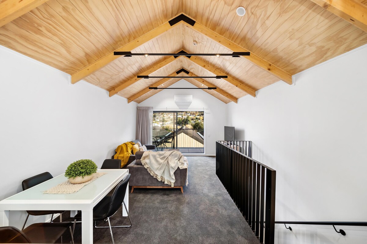 An open-plan living area is presented with a high, wooden beam ceiling, enhancing the spacious feel. A dining table is positioned beside a cozy seating arrangement, with large windows allowing natural light to fill the space and offering views of the surroundings.