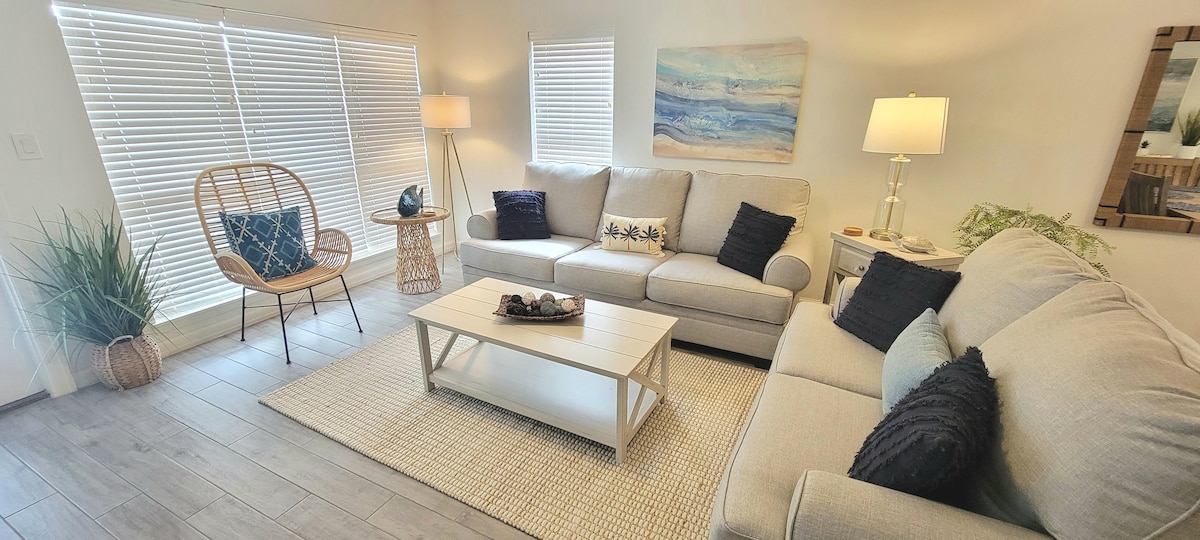 A bright living room is centered around a light wood coffee table, with a large sectional sofa offering comfortable seating. Natural light filters through sleek blinds, highlighting the neutral tones of the decor. A rattan chair and potted plants add texture to the space.