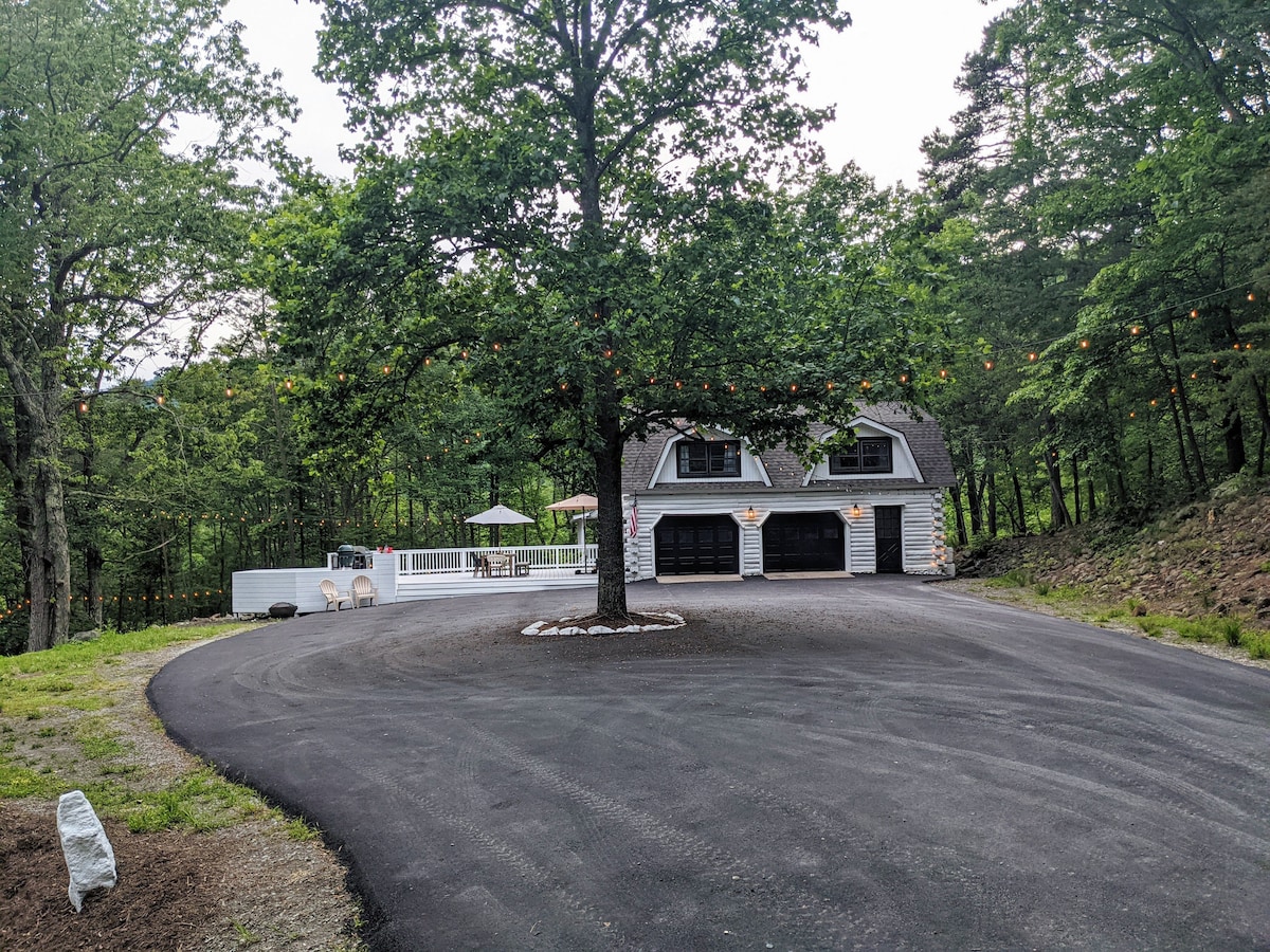 A spacious circular driveway is surrounded by greenery, leading to a two-car garage with decorative windows. A large tree provides shade, and patio furniture is visible on a raised deck, creating an inviting outdoor area for relaxation.