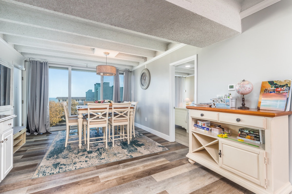 A dining area features a wooden table surrounded by six chairs, complemented by natural light coming through large sliding doors. Light grey curtains frame the view of the balcony, which includes a glimpse of the ocean. A side cabinet holds various games and books.