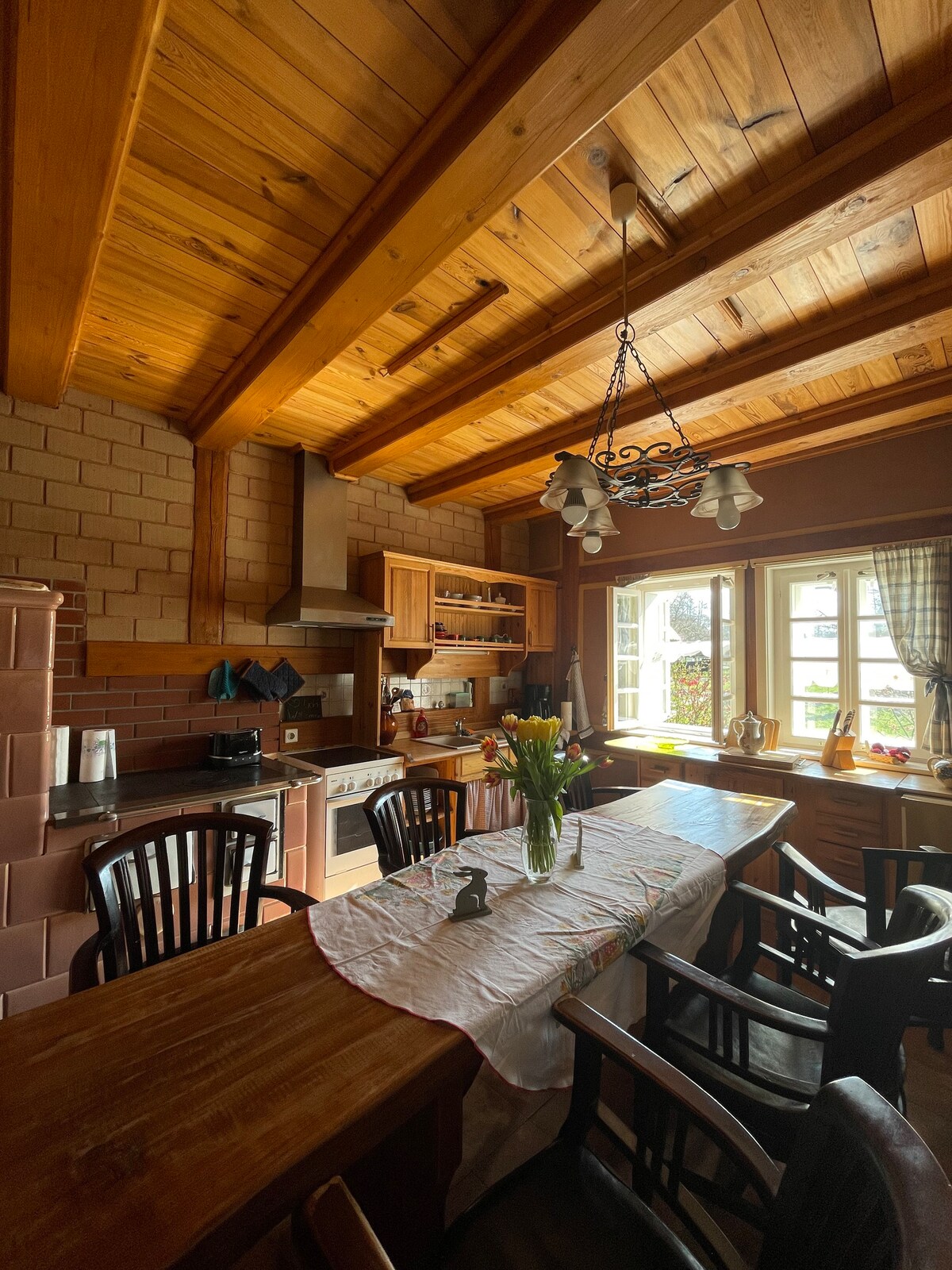 A rustic kitchen is showcased with wooden beams overhead and a long table set for dining. The space features a variety of cabinets and a stove, along with large windows allowing natural light to illuminate fresh flowers placed in a vase.