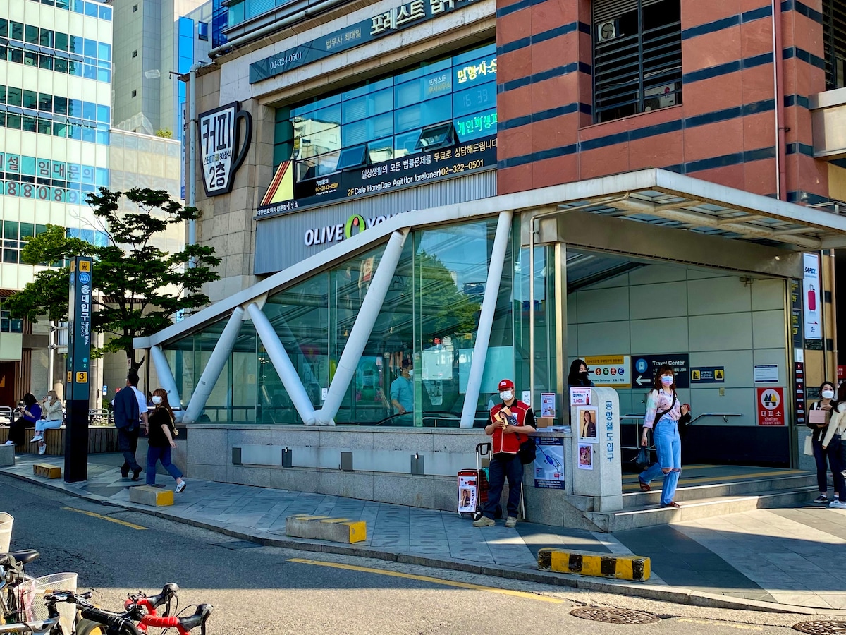 The image shows the entrance to a subway station, featuring a modern glass structure supported by white beams. Pedestrians can be seen entering and exiting, while various signage is visible. The surrounding area includes trees and buildings, indicating a lively urban environment.
