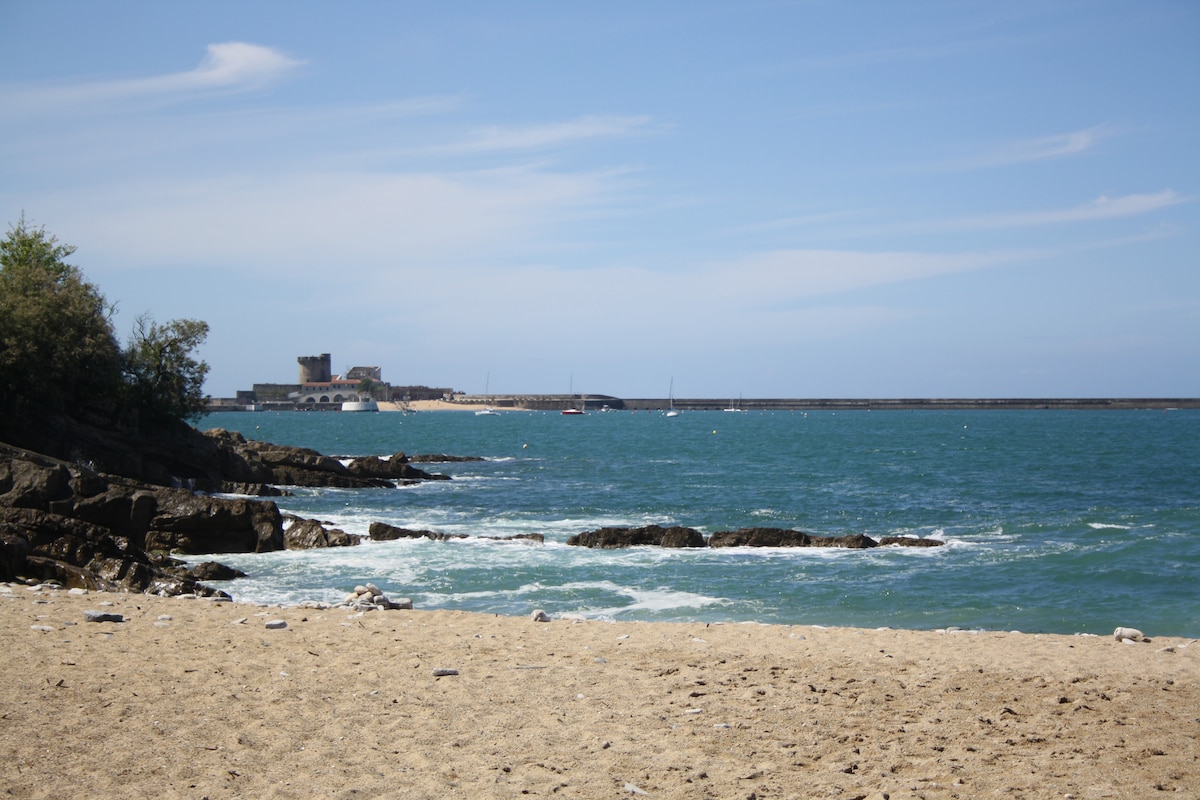 The beach presents a sandy shoreline beside calm blue waters, with gentle waves lapping against it. In the distance, a fortified structure is visible along the coastline, complemented by boats anchored in the harbor. A clear sky provides a serene backdrop.