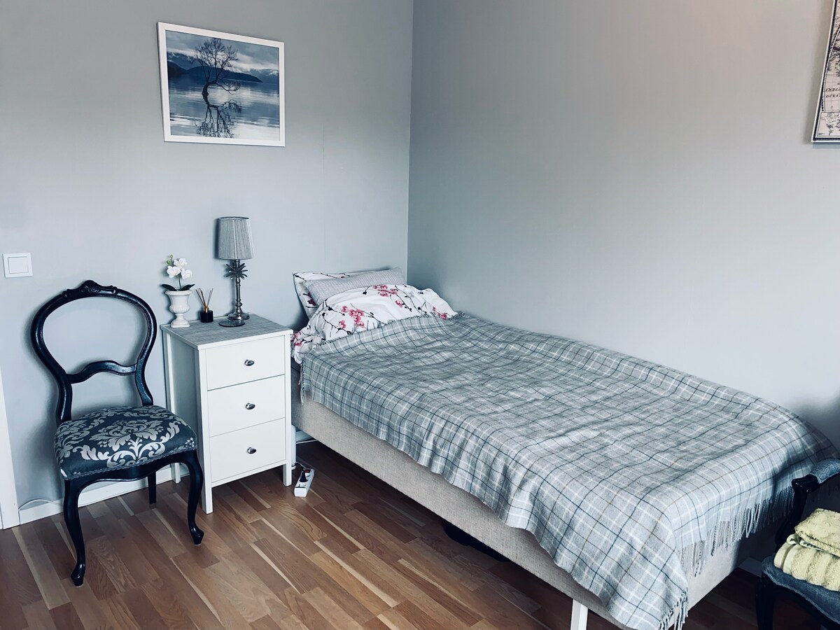 A clean sleeping area is presented with a single bed covered by a gray plaid blanket. A decorative chair stands beside a nightstand that holds a lamp and a small floral arrangement. Light wood flooring complements the soft gray walls.
