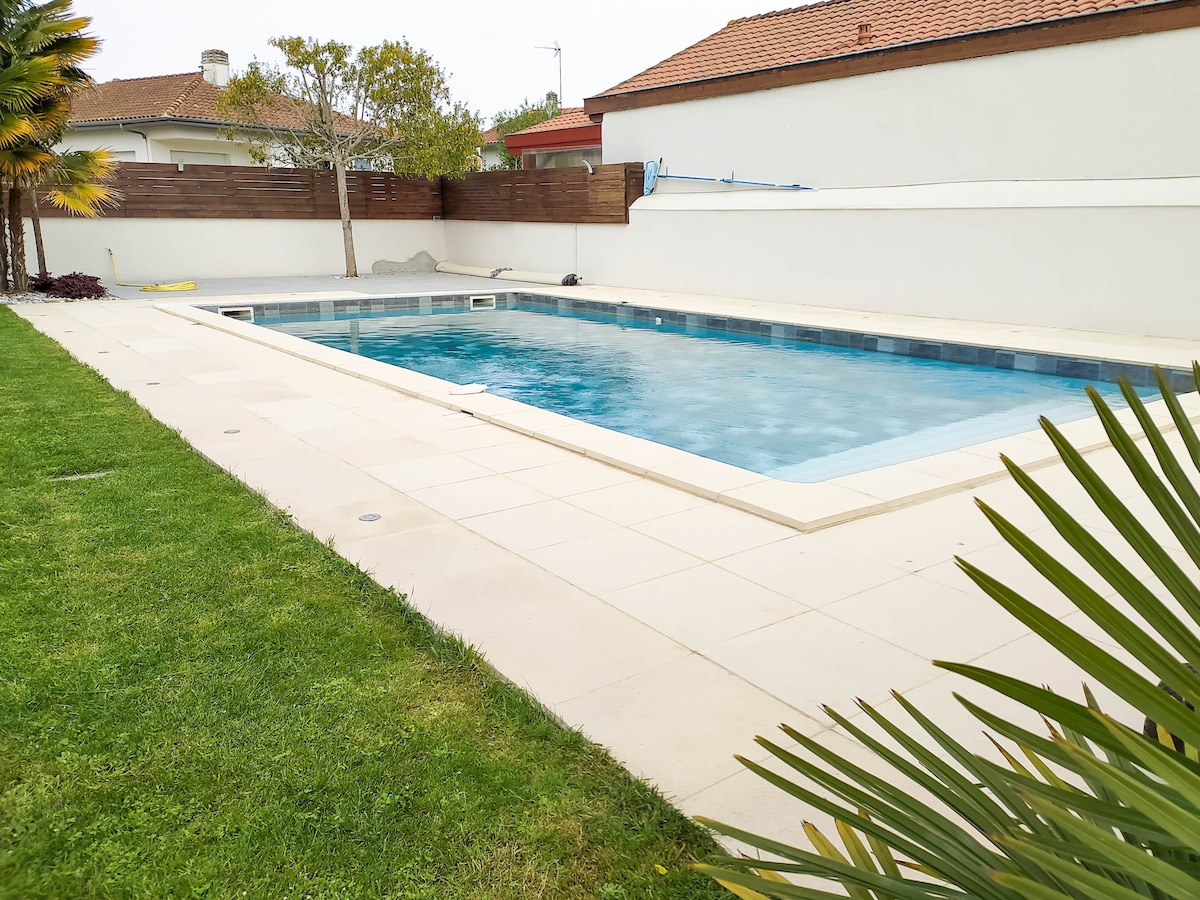 A private, heated swimming pool is set within a landscaped garden. The spacious pool features clear water and is bordered by tiled areas for sunbathing. Soft green grass surrounds the pool, providing a pleasant outdoor space for relaxation.