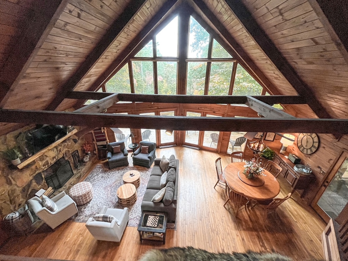 An open-concept living area showcases a high ceiling with exposed wooden beams and large floor-to-ceiling windows. Comfortable seating arrangements are present around a rustic coffee table, with a dining area featuring a round table and chairs nearby. Natural light illuminates the space, highlighting the wooden flooring.