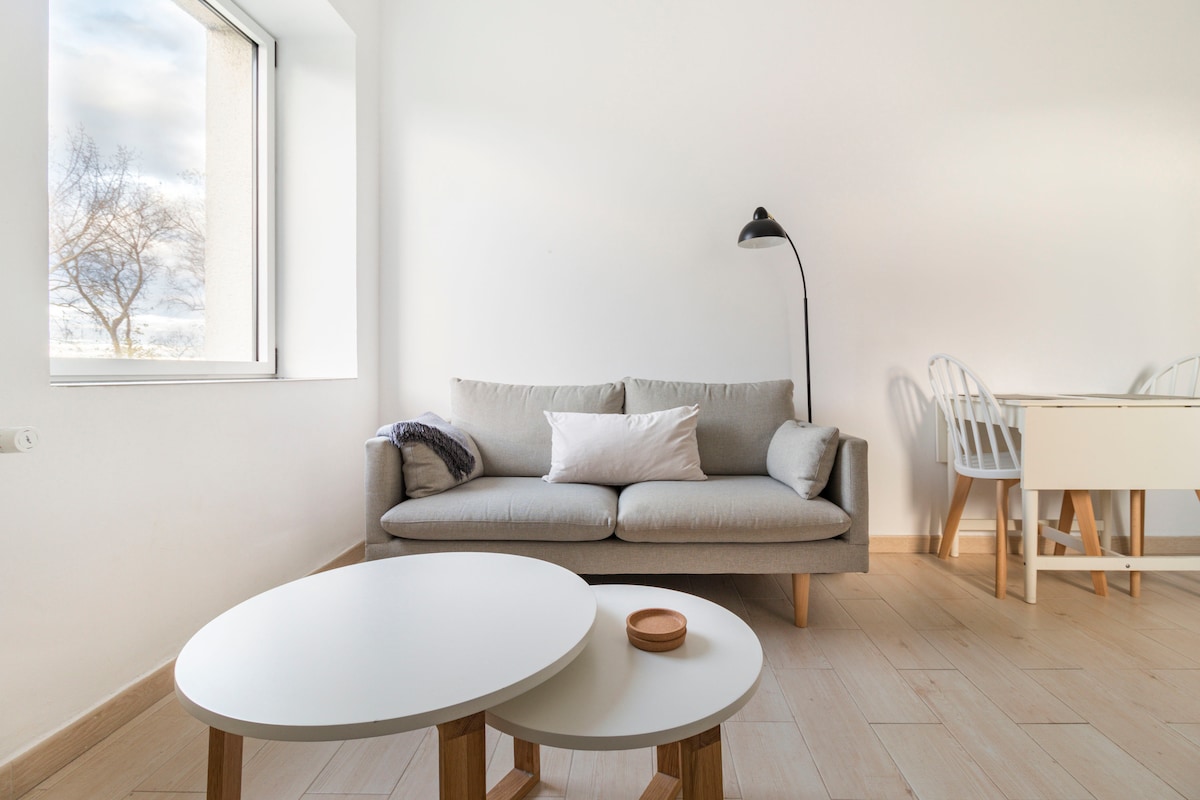 A light-filled living area features a comfortable gray sofa with a decorative pillow. Two round white coffee tables are positioned in front of the sofa. A floor lamp provides illumination. In the background, a dining space is visible, with a white table and chairs near a window.