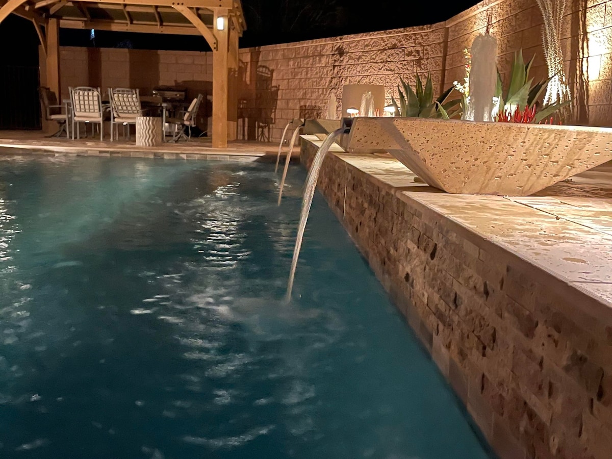 A tranquil pool area is featured, highlighted by gentle water cascading from a decorative stone feature into the clear blue water. Comfortable seating is arranged nearby under a wooden structure, surrounded by softly illuminated plants, creating a serene outdoor environment.