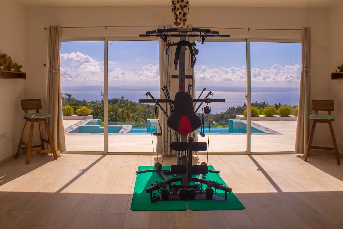 A functional workout space is equipped with exercise machinery, positioned on a green mat. Large sliding glass doors provide a panoramic view of the outdoor pool and surrounding landscape, allowing natural light to fill the area.