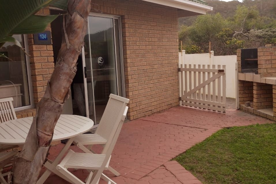 An outdoor area is seen, featuring a small enclosed garden with a circular table and white chairs on a brick patio. A gate leads to the garden, and a braai area is positioned nearby against a brick wall.
