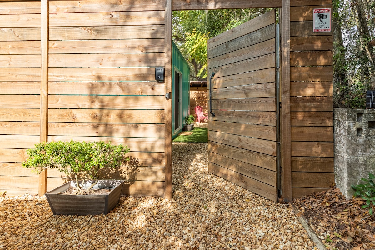 A wooden gate is open to reveal a gravel pathway leading into a lush garden area. Flanking the path is a neatly arranged planter with greenery, complemented by a backdrop of rustic wooden fencing and a vibrant green container home nestled within the trees.