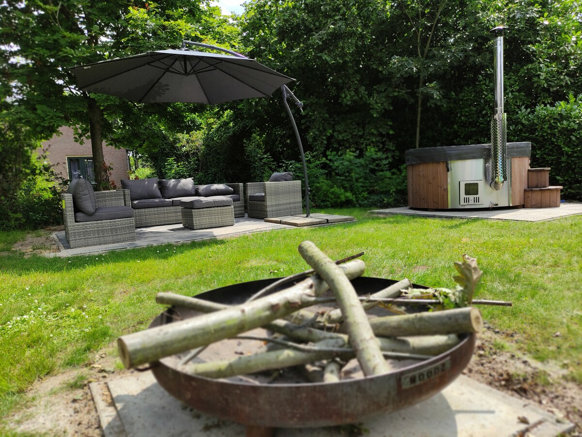An outdoor seating area features a shaded lounge set and a wood-burning hot tub, surrounded by lush greenery. A fire pit, filled with logs, is situated in the foreground, providing a cozy space for gatherings.