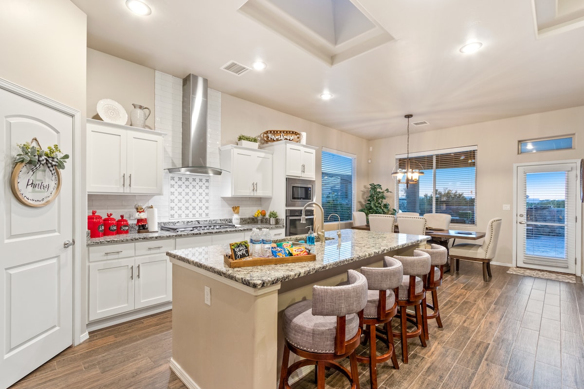 A modern kitchen features light cabinetry and a spacious island with seating for six. Stainless steel appliances and decorative accents are arranged on the countertops. Large windows provide natural light and a view of the outdoor space, enhancing the inviting atmosphere.