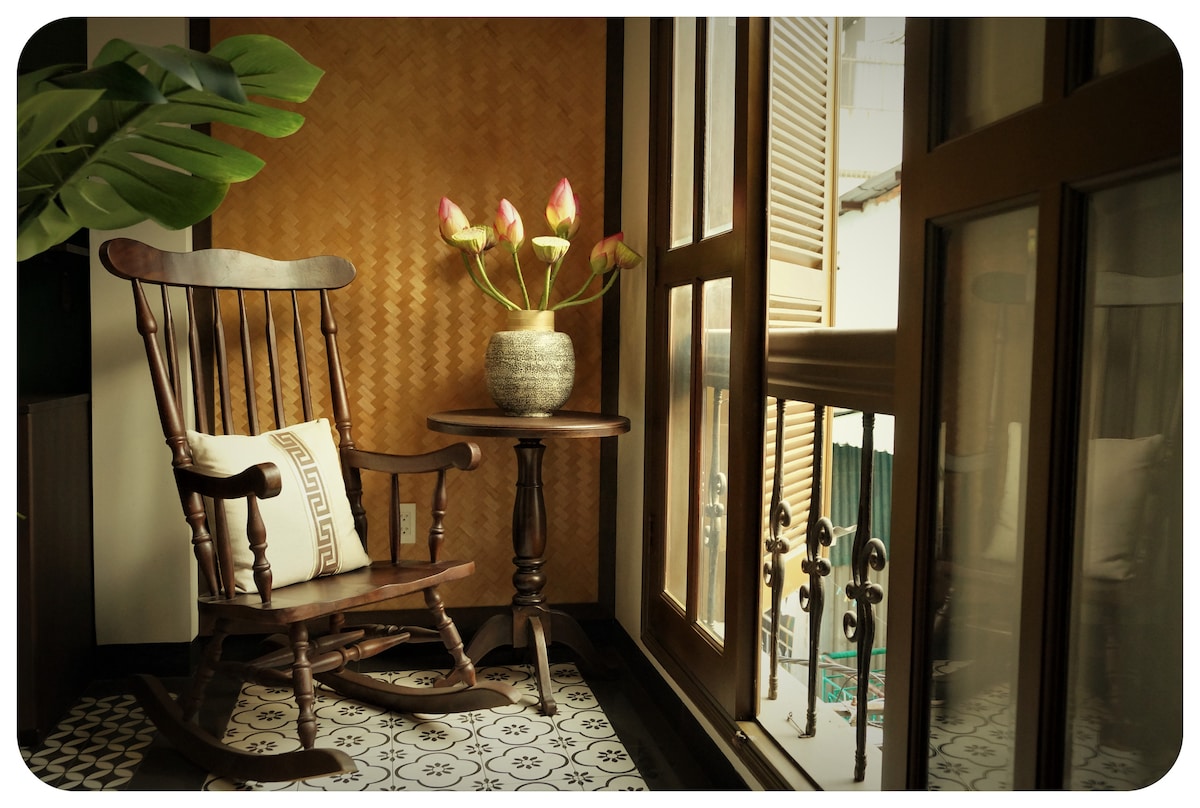 A vintage rocking chair is positioned next to a large window, bringing in natural light. A small wooden table holds a vase with pink lotus flowers. The patterned tiled floor and textured wall create a warm, inviting space.