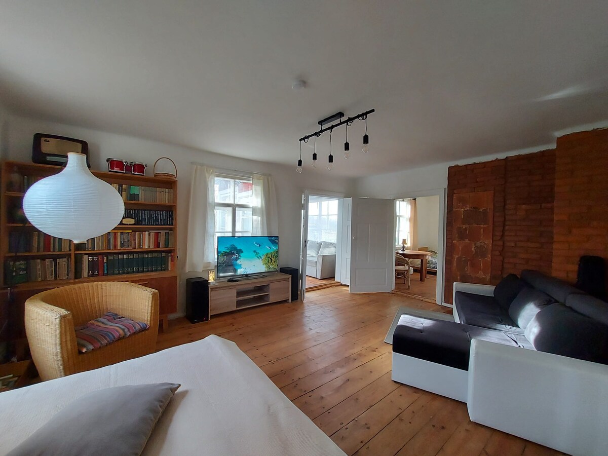 A spacious living area features a soft sofa and a modern television. Natural light enters through large windows, illuminating the wooden floor. A wicker chair adds to the seating options, and a bookshelf displays an array of books, enhancing the room's inviting character.