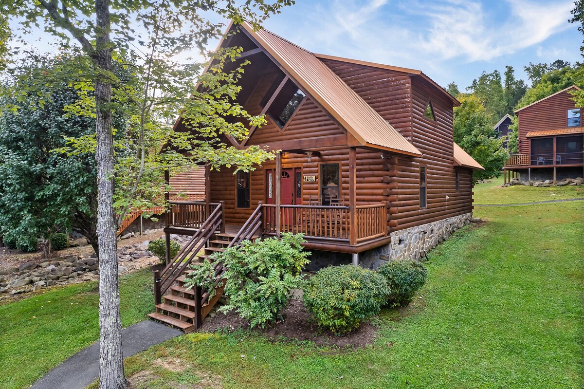 The cabin features a charming wooden exterior with a front porch and multiple steps leading up to the entrance. Lush green landscaping surrounds the property, enhancing its natural setting. The structure showcases a distinctive roof design and large windows that invite light into the space.