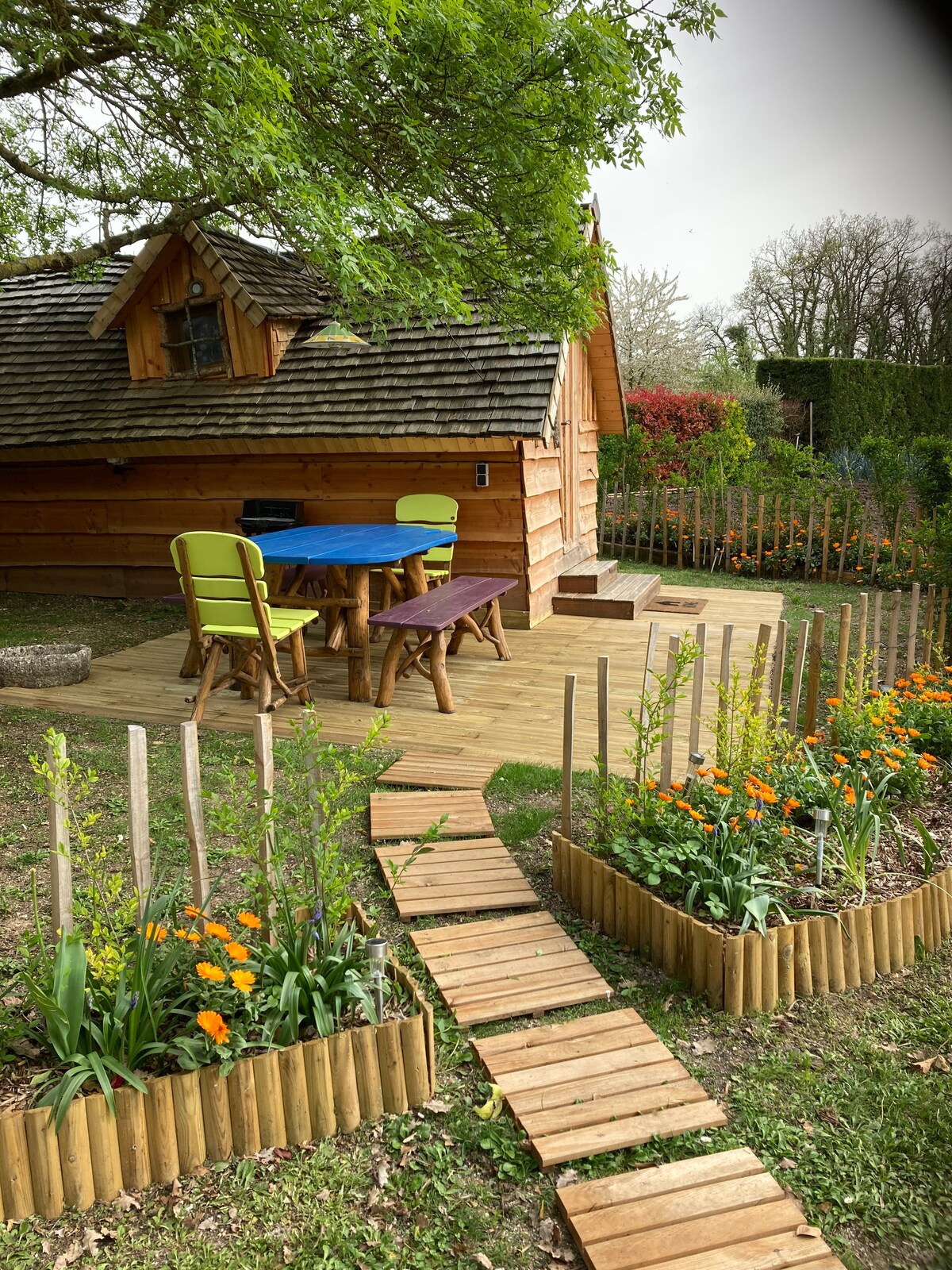 A charming wooden cabin is surrounded by lush greenery and colorful flowers. A wooden deck hosts a table with chairs in bright colors, inviting outdoor relaxation. A pathway made of wooden planks leads through landscaped flower beds, enhancing the tranquil garden setting.