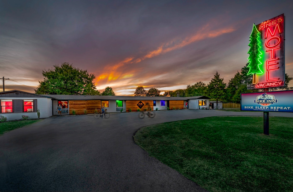 The exterior of The Bike Inn is captured at dusk, featuring a winding driveway leading to a single-story building. A vibrant neon sign illuminates the property, while a colorful sunset casts a warm glow over the surrounding trees. Bicyclists can be seen in the vicinity.